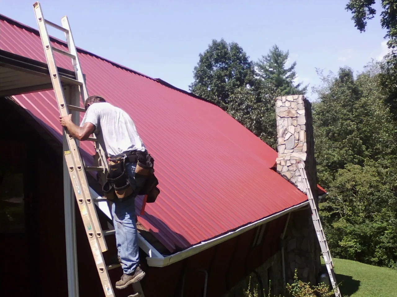 Roofer on a ladder beside a red metal roof and stone chimney — Soffit & Fascia Repair by Roofing Brothers in Gordon